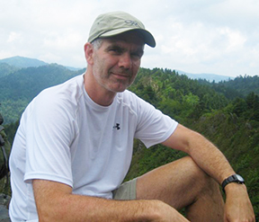 Roger Traylor resting on a hike with forested hills behind him.