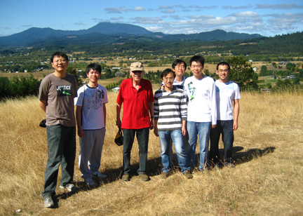 Gabor Temes posing with his graduate students on a dry, grassy hill with lush rolling hills and a valley in the background.