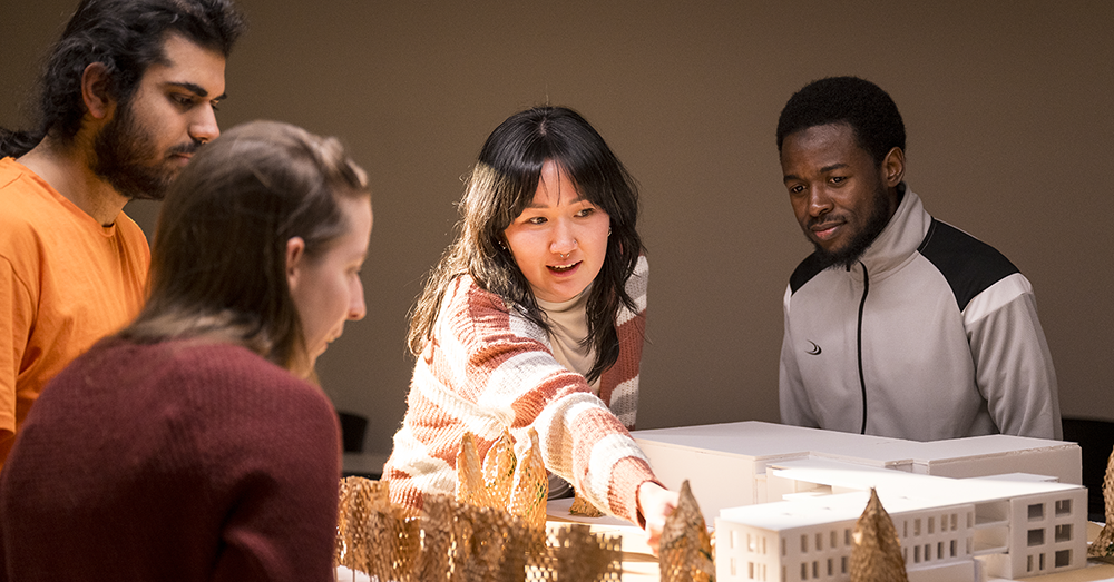 A group of engineering students stands over a table with a scale model of a building and trees, a central woman reaches over pointing at an architectural detail.