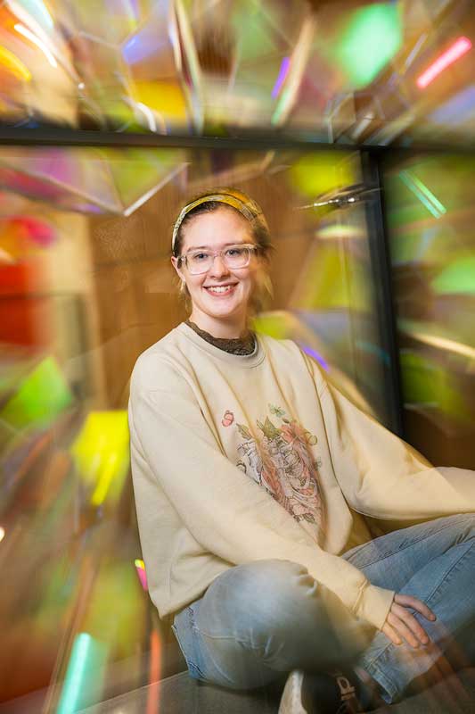 A student sitting cross-legged on a table in a classroom with colors reflecting from an art installation appearing in different shapes around the student’s face.