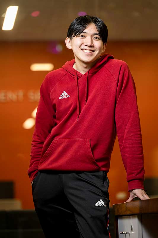 A student wearing a sweatshirt leaning against a table and smiling at the camera while standing in a classroom.