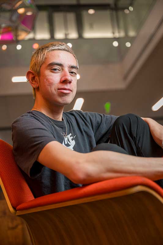 A student sitting in a chair looking down at the camera in the lobby of Johnson Hall with an art installation above.