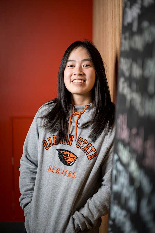 A student wearing a Oregon State University sweatshirt is leaning against a chalkboard with writing looking at the camera.