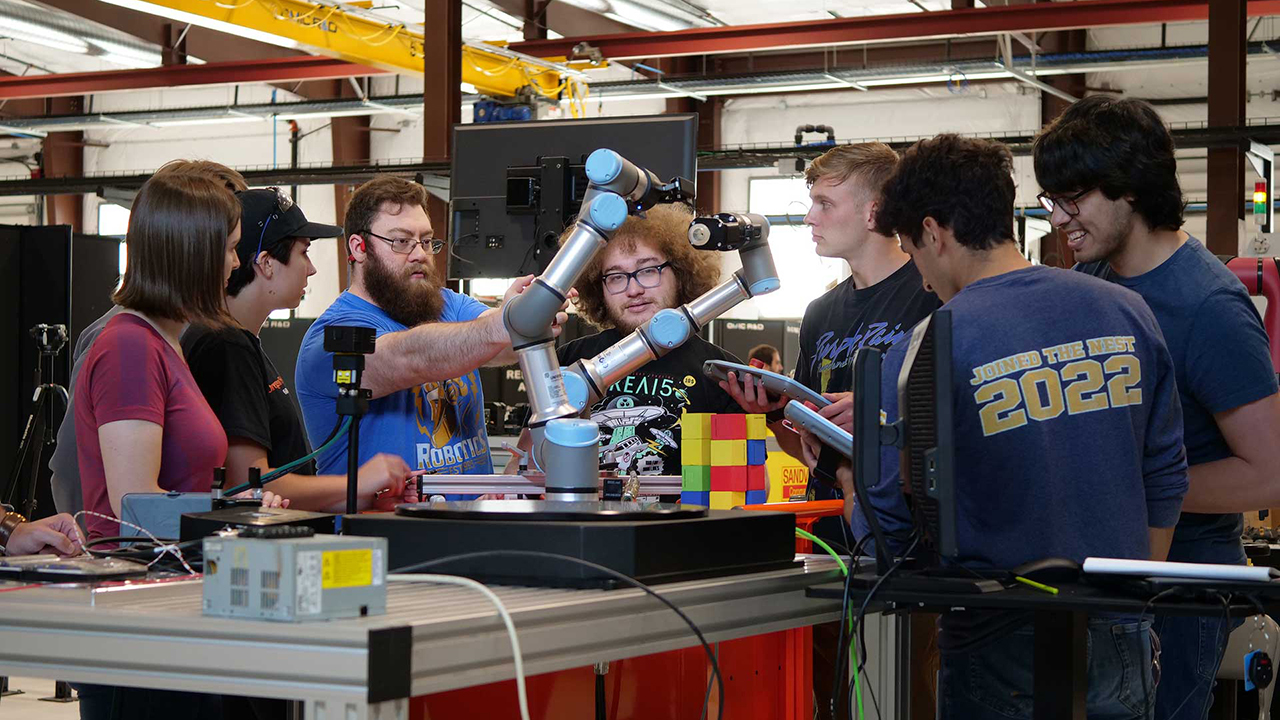 Eight students standing around a workstation at a lab with one student adjusting a robotic arm.