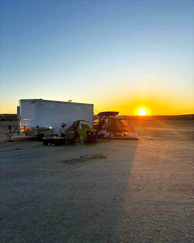 Four people sitting in chairs around two tents and a trailer in desert with the sun setting in the background.