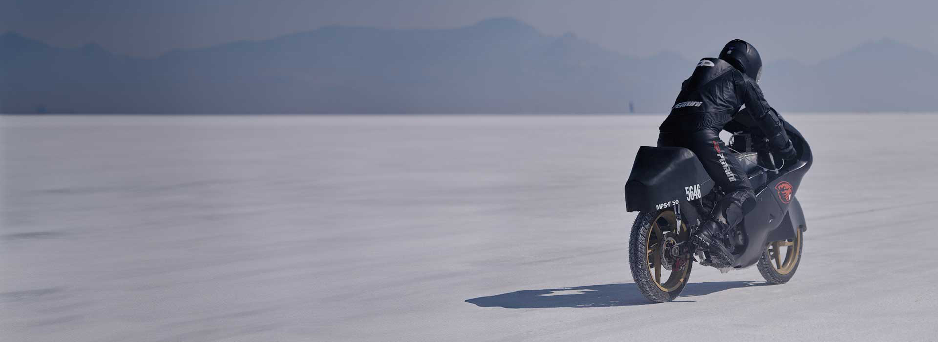 A motorcyclist riding a 50cc motorcycle across desert sand with mountains in the distance.