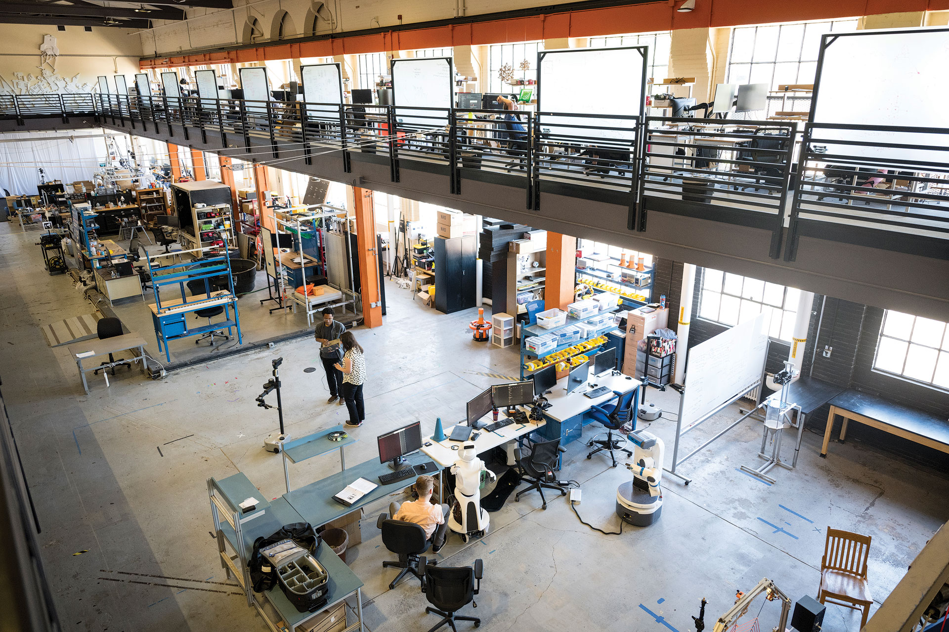 Two researchers standing in the center of a large open-room, multi-story research facility with robotics equipment and desks surrounding them. 