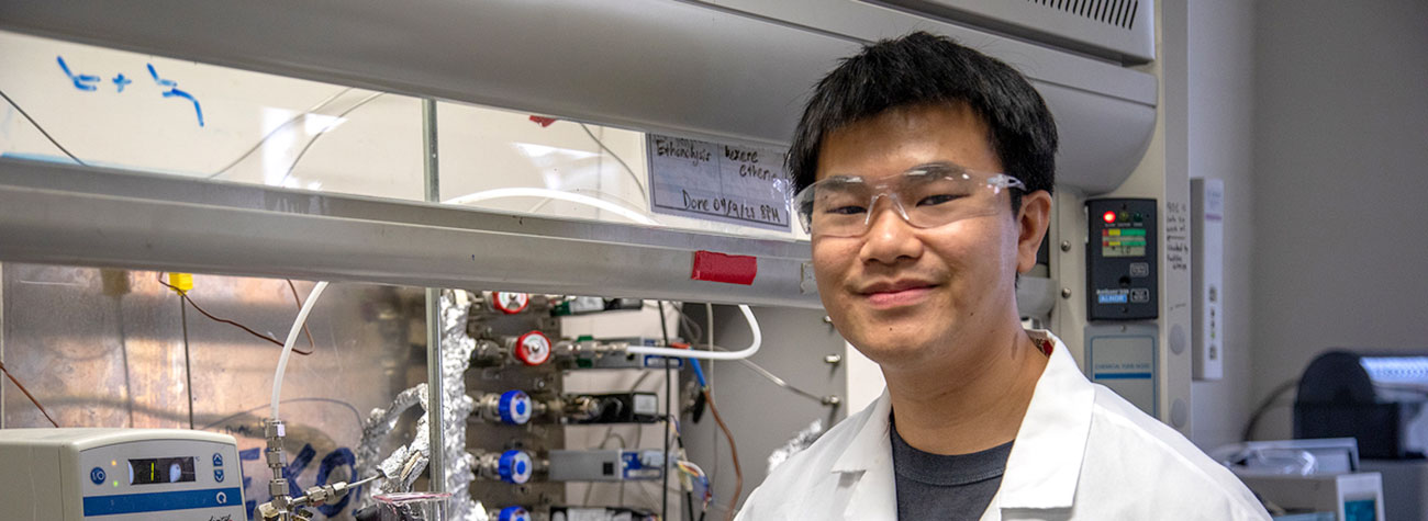 A person wearing a lab coat and eye protection stands in front of equipment in a lab facing the camera.