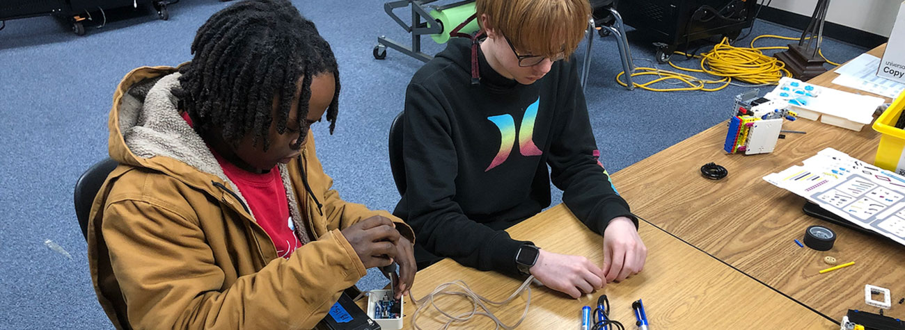 Two students sit at a table and adjust robotics equipment. 
