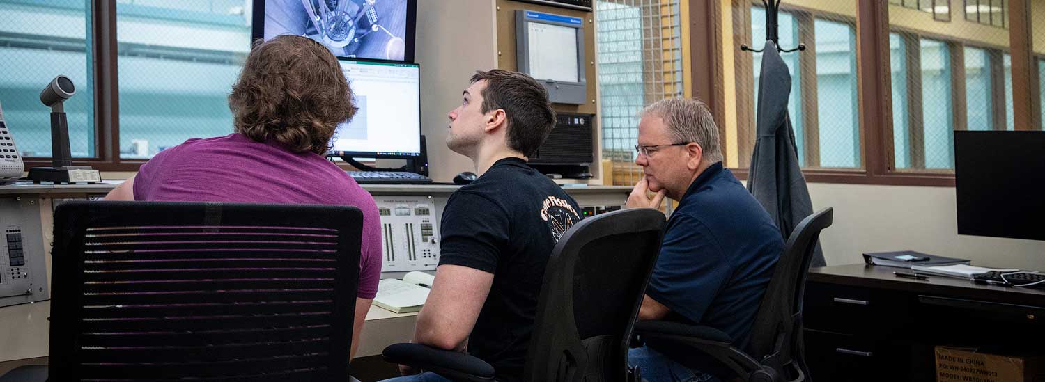 Three people sit in chairs and look at control systems of a nuclear reactor.