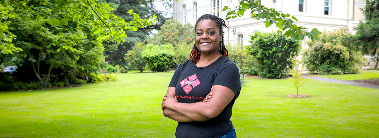 A person stands outside and smiles toward the camera with green grass and a historic building in the background.