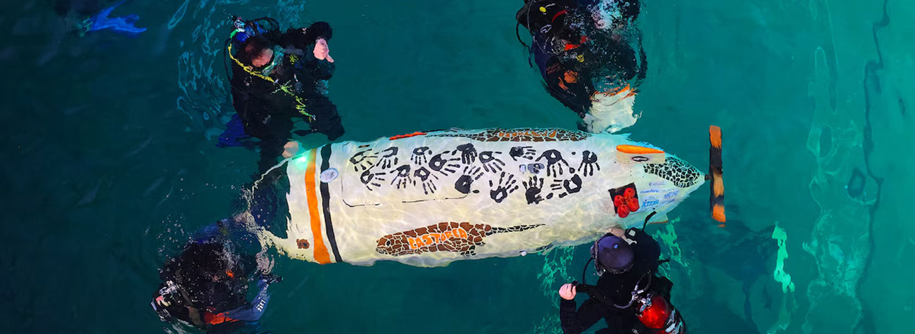 Four people in scuba diving gear float on the surface of water and touch a small submarine with the word Castorea painted on its side.
