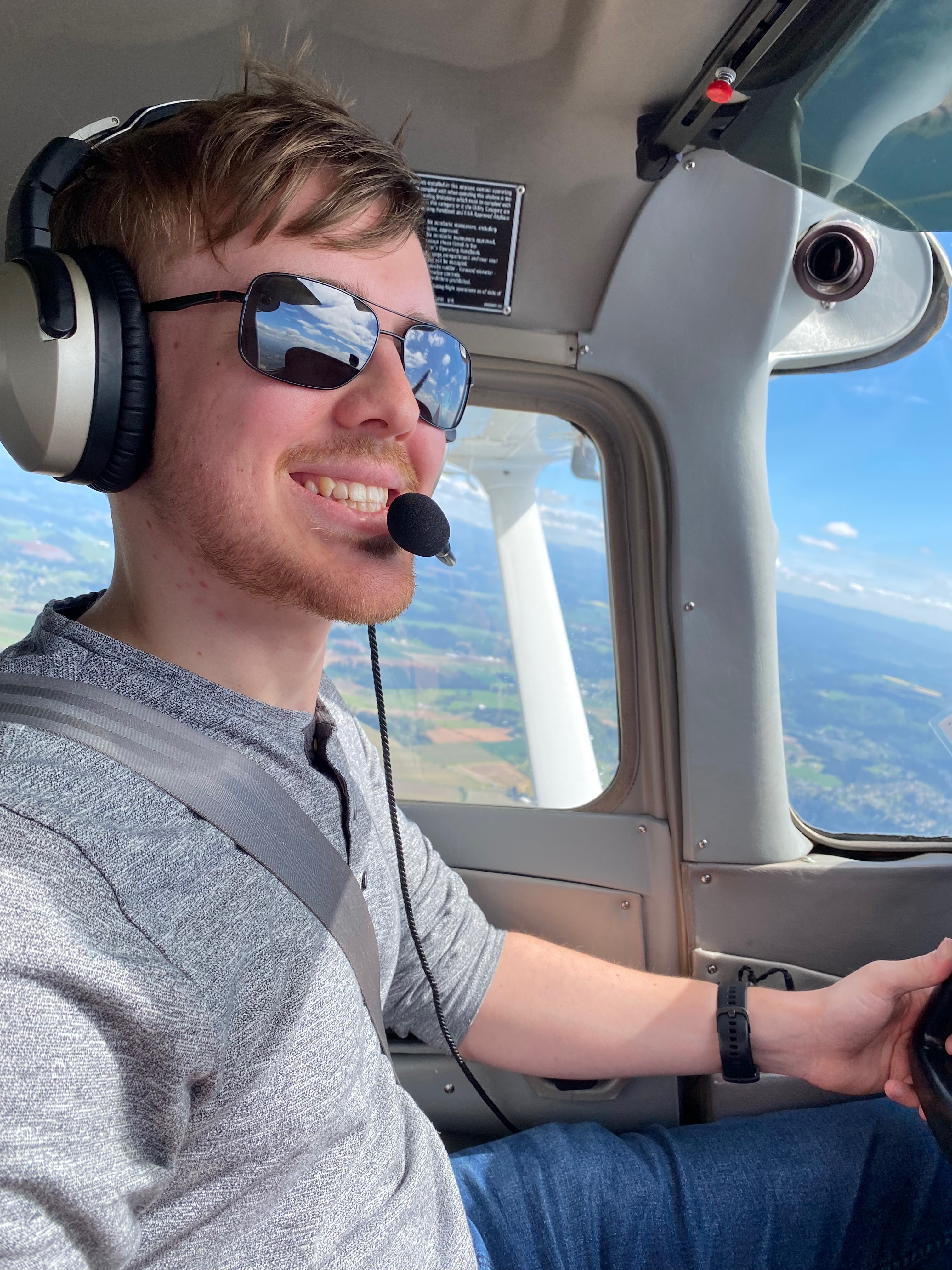 A man sitting in the cockpit of an airplane with headphones and sunglasses, smiling at the camera.