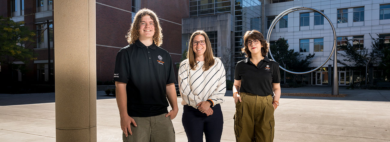 Three people stand outdoors on a campus with circular sculpture in the background and smile toward the camera. 