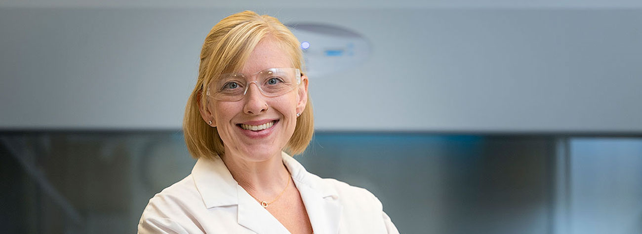 A person stands in a lab wearing PPE and smiles toward the camera.