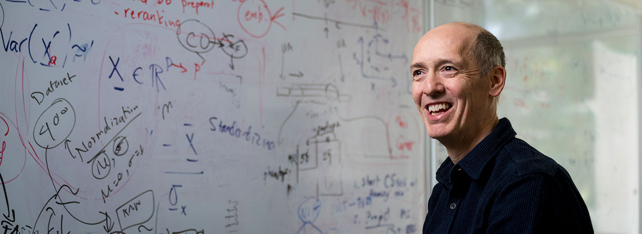 A person stands in front of a whiteboard with figures on it and smiles toward a subject of camera.