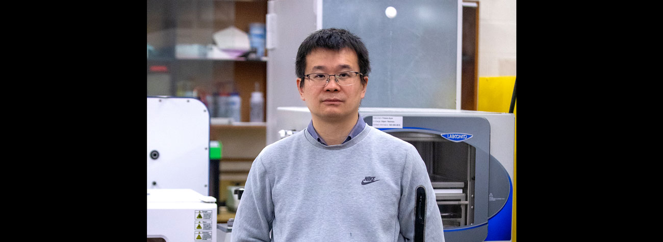 A person wears a light grey sweatshirt and stands in front of industrial equipment in a lab and looks toward the camera.