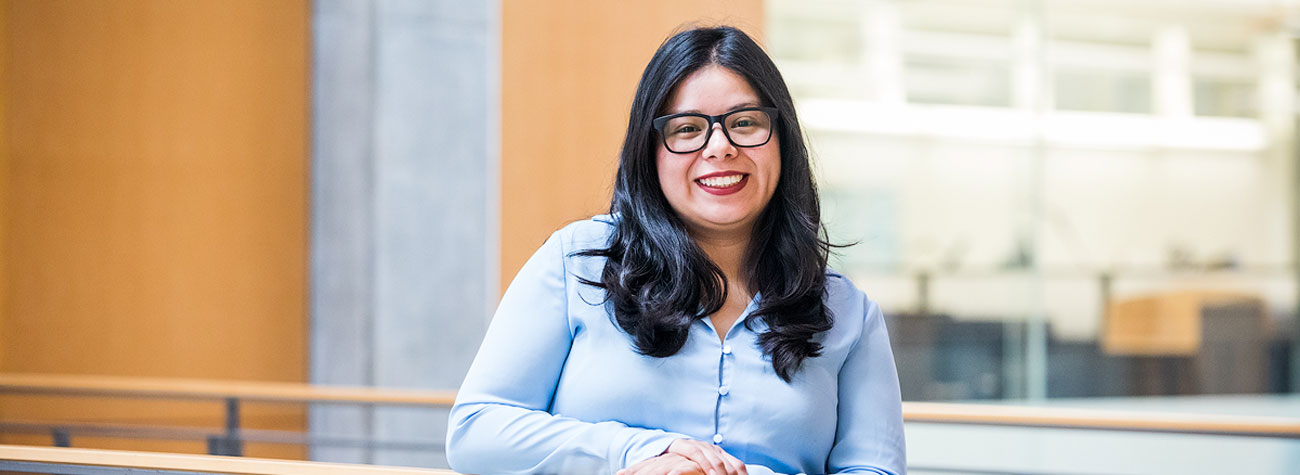 A person stands indoors near a wooden railing with a modern office background featuring glass panels and neutral-colored walls.