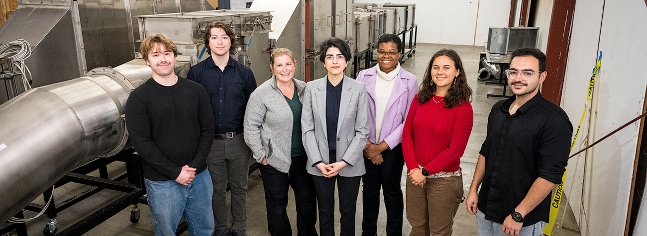 Seven people stand in a lab next to cylindrical equipment and look up toward the camera.