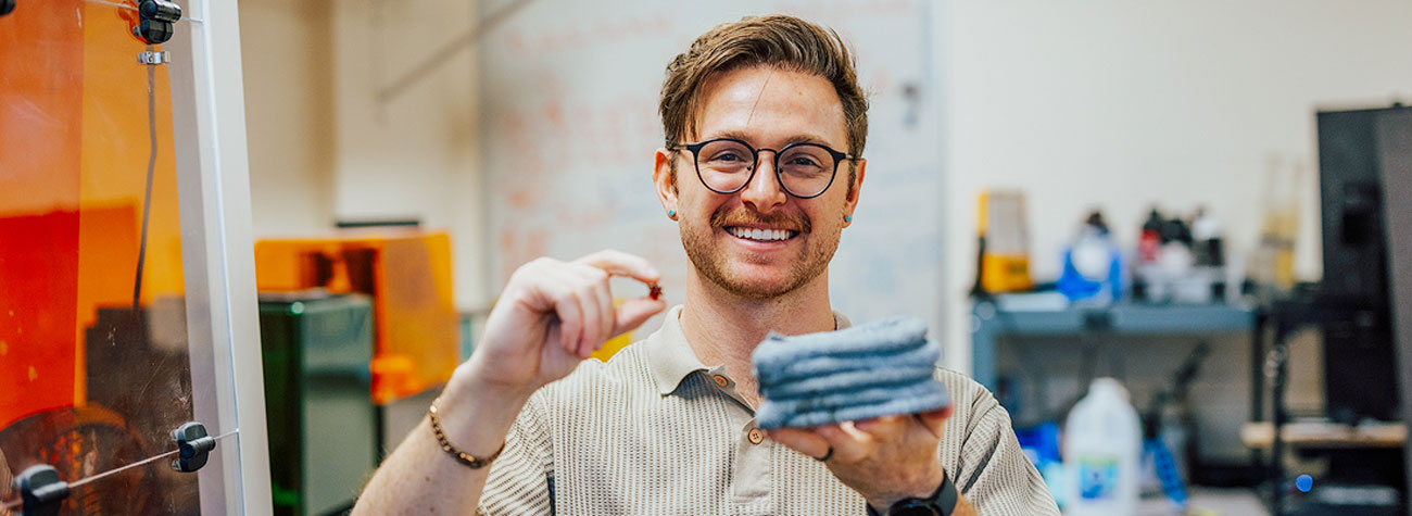 A person stands in a lab and holds a small item in their right hand and a stack of flat blue gray materials in their left hand and smiles toward the camera.