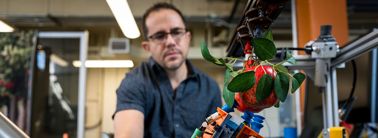 A person in a lab setting looks down at a robotic arm next to an apple. 