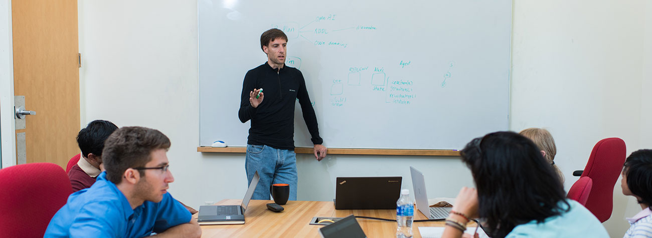 A person stands at the front of a conference room, pointing to a whiteboard covered with handwritten diagrams and notes, while several people sit around a table with laptops and notebooks, listening and discussing.
