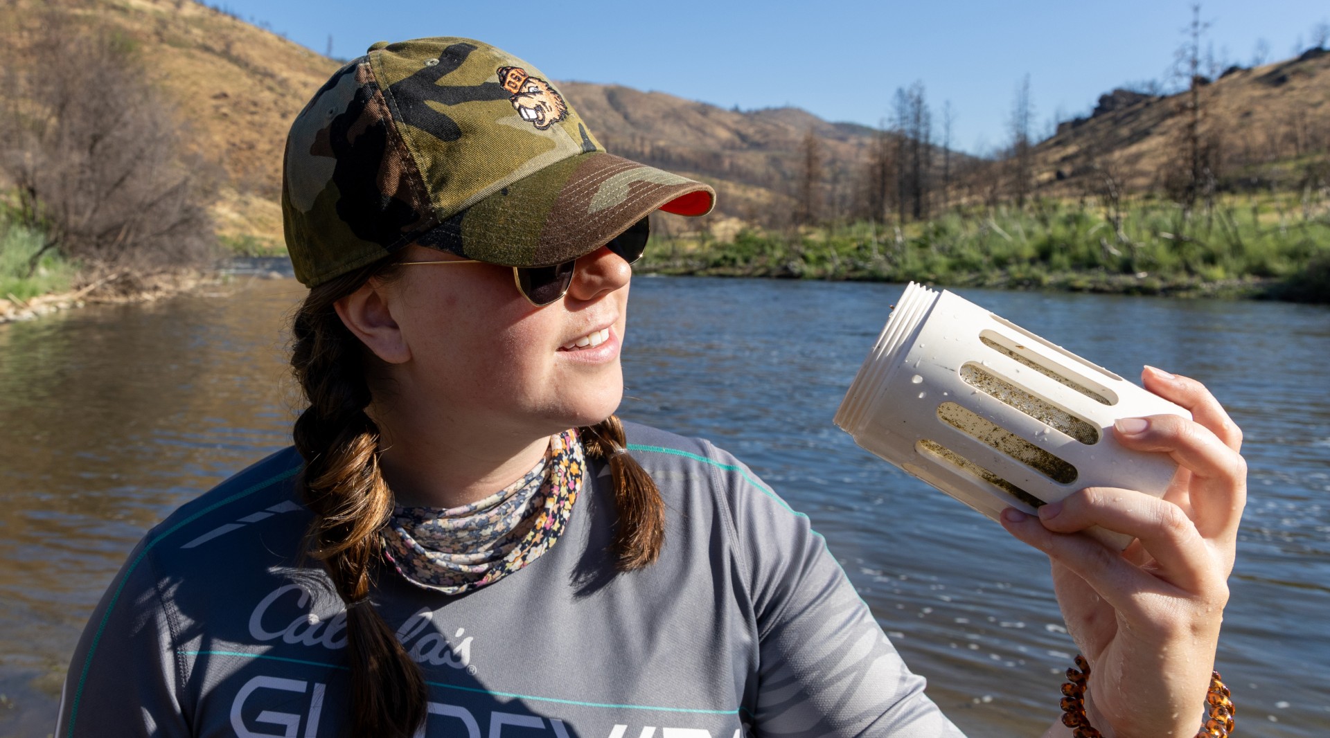 Whitney Packard in a baseball cap and sunglasses examines a white container containing particle samples collected from the Klamath River.