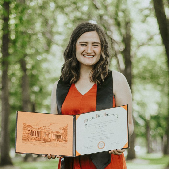 Tiana Tonge holding a diploma open, standing outside with trees and vegetation.