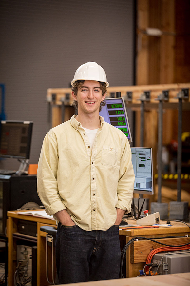 Dylan Goff wears a hard hat and stands in front of digital screens in the Emmerson Lab, a large timber engineering facility, and smiles toward the camera.