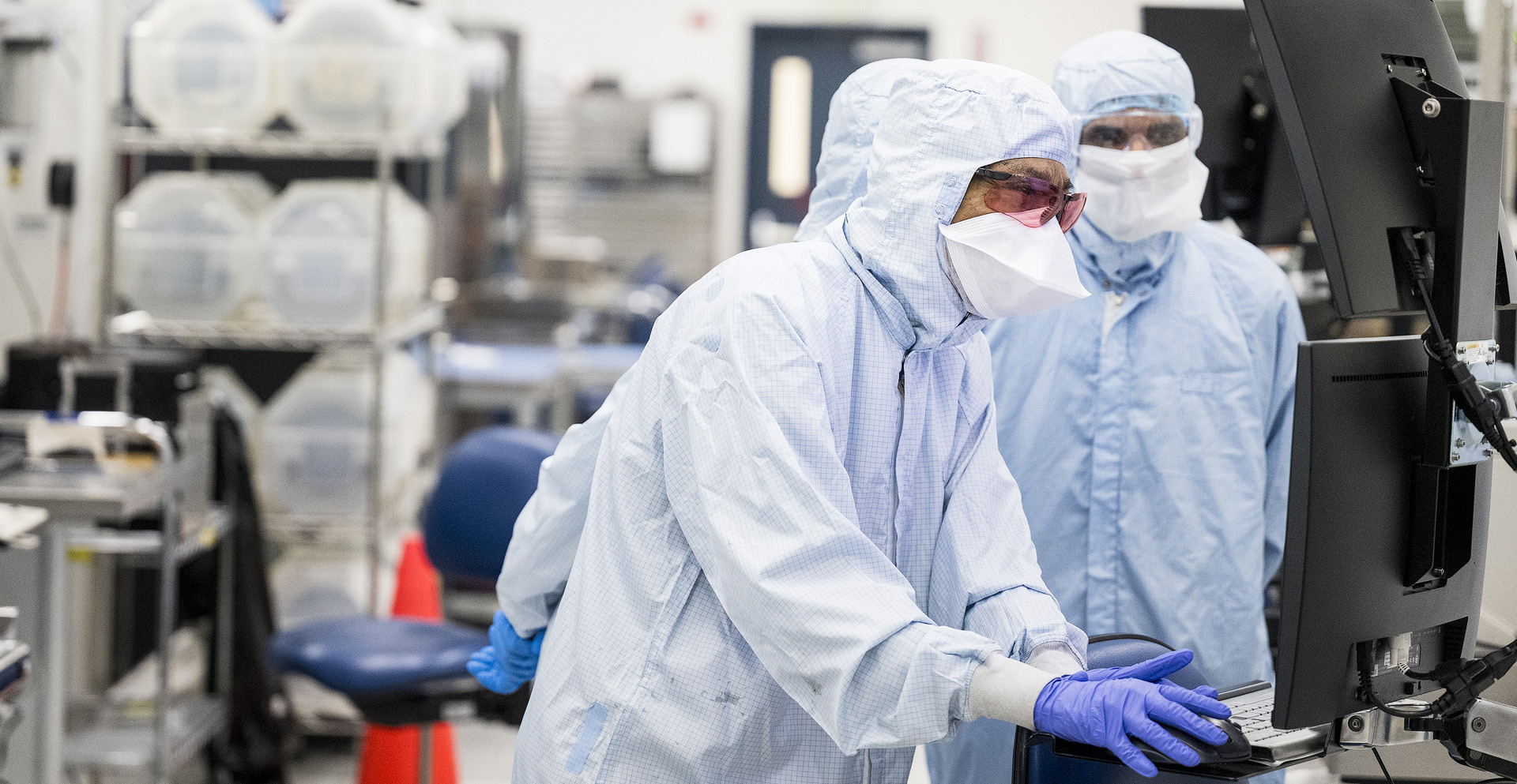 Two researchers in cleanroom suits looking at monitors.