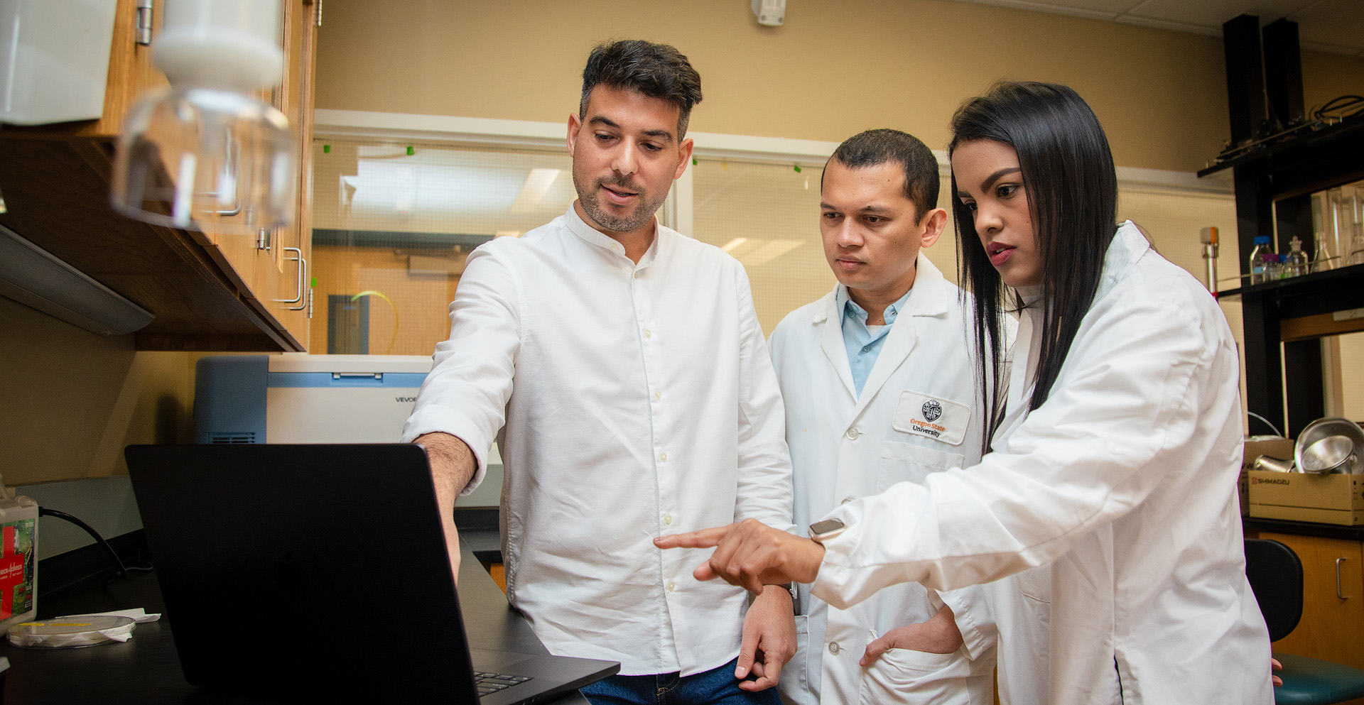 Three workers in lab coats looking at a laptop.