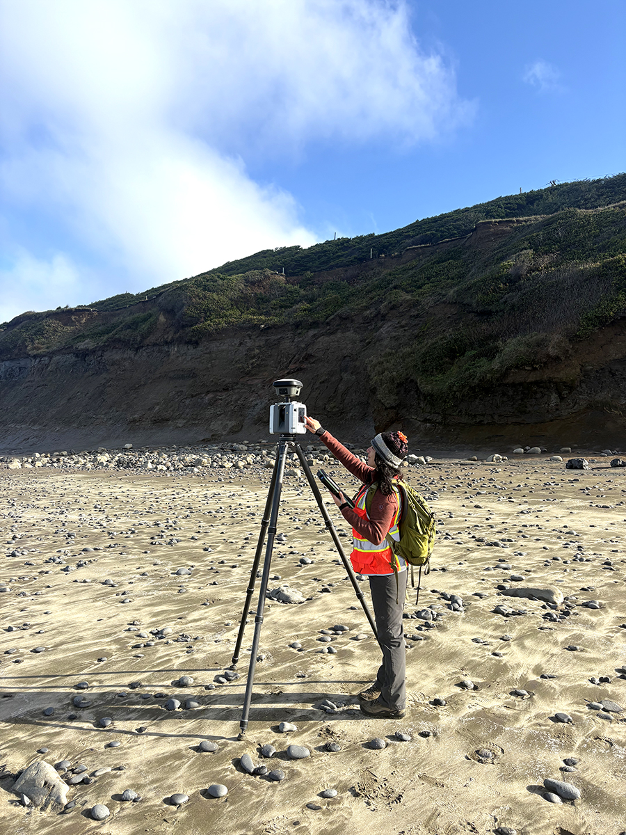 Heather Maran stands next to a tripod-mounted lidar scanner on a beach below an eroding coastal bluff.