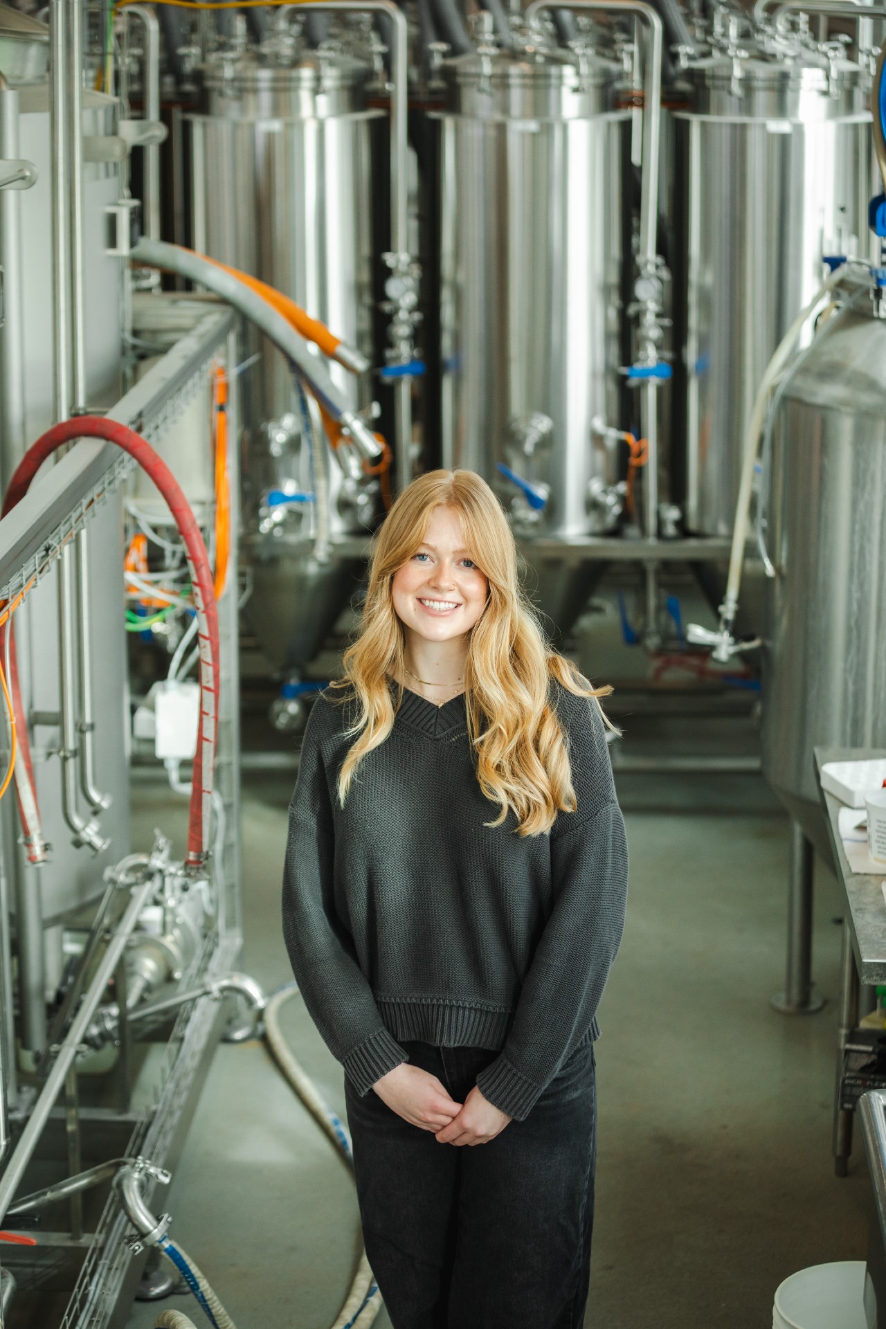 Undergraduate engineering student, Leah Byrne, stands in front of tall stainless-steel industrial tanks and pipes with a production facility in the background.