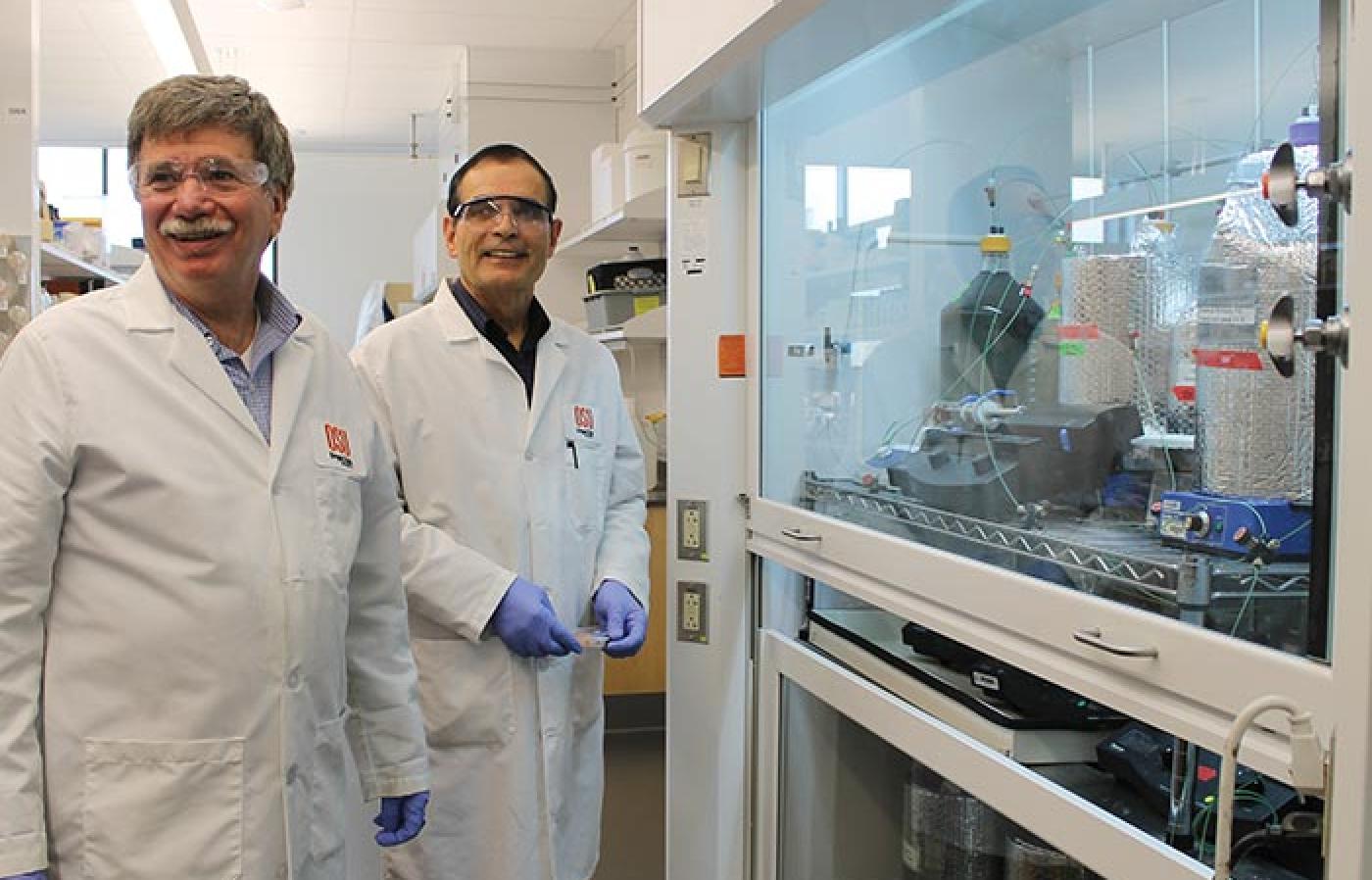 Two faculty member wearing lab coats in front of a protected glass with scientific materials inside it.