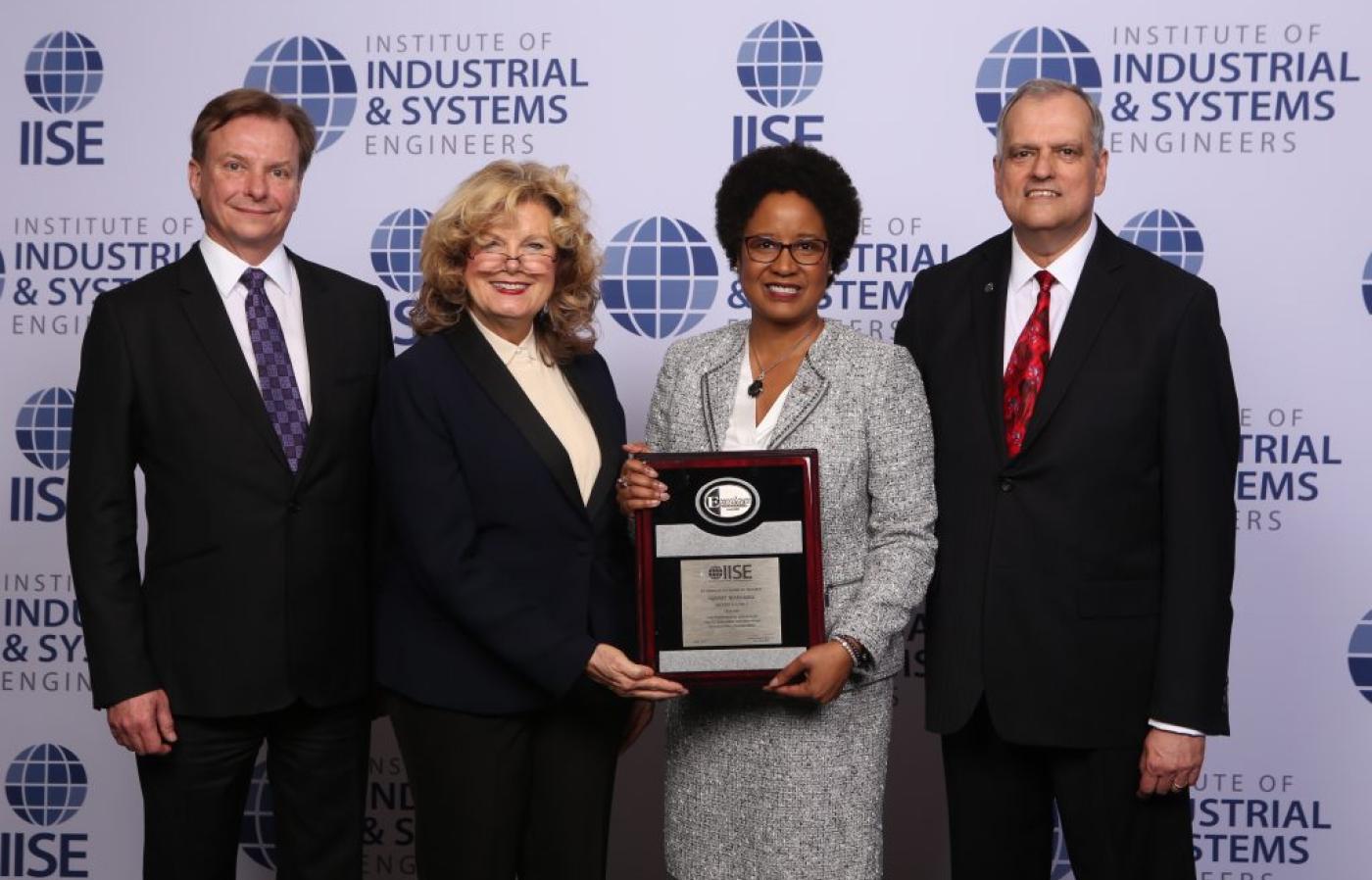 Four professionals stand before an IISE backdrop as one holds a framed award during a formal recognition event.
