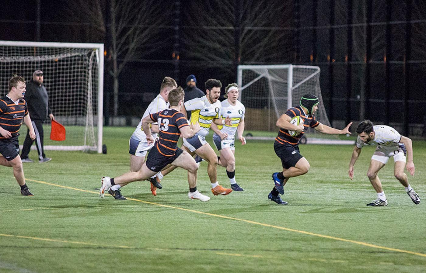 Rugby players sprint toward the try line under stadium lights as a ball carrier evades defenders on a turf field.