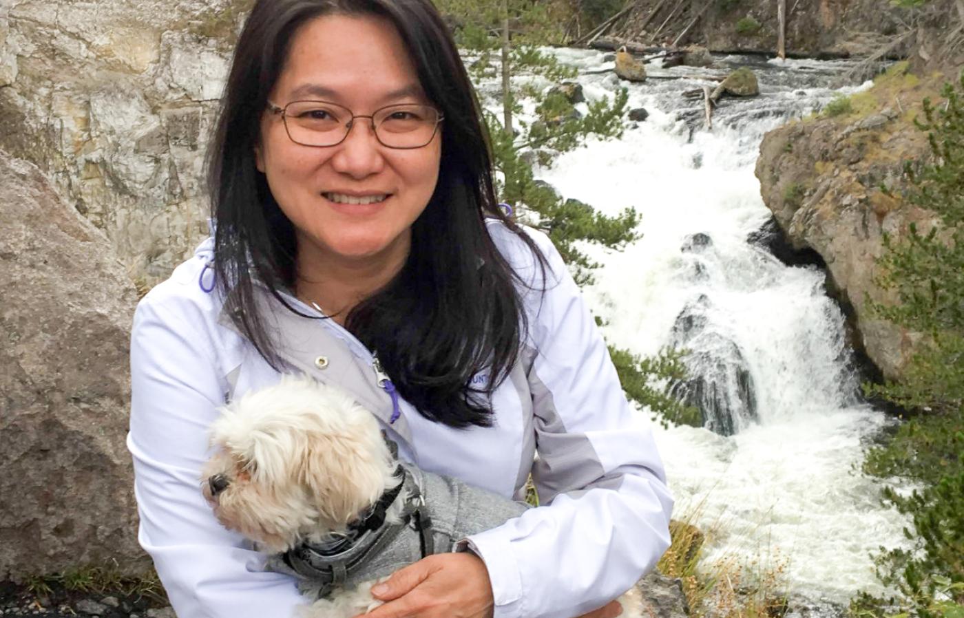 Leanne Lai holding a small white dog in front of a rapid creek waterfall.