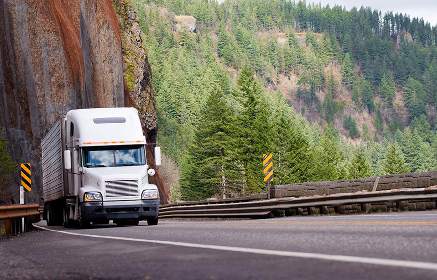 A white semi-truck driving on a mountainous road with trees and a rocky cliff.