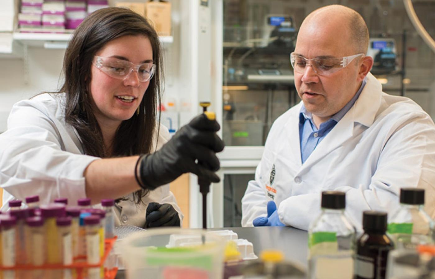 Two people wearing lab attire while working together in a lab.