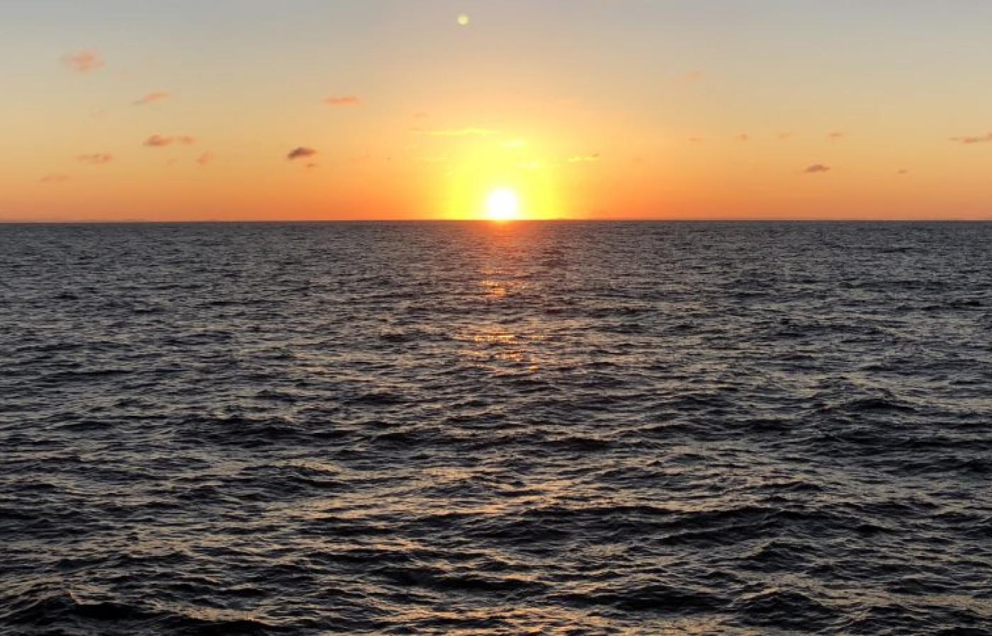 The sun sets on the horizon, casting warm orange light across gently rippling ocean waves beneath a clear sky.