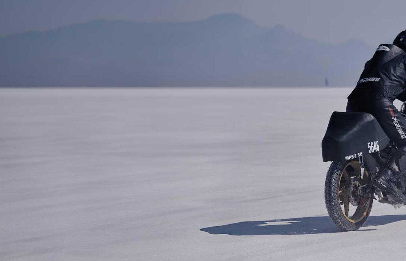 A motorcyclist riding a 50cc motorcycle across desert sand with mountains in the distance.