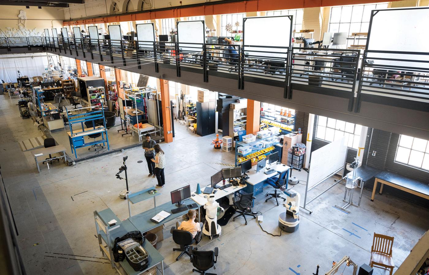 Two researchers standing in the center of a large open-room, multi-story research facility with robotics equipment and desks surrounding them. 