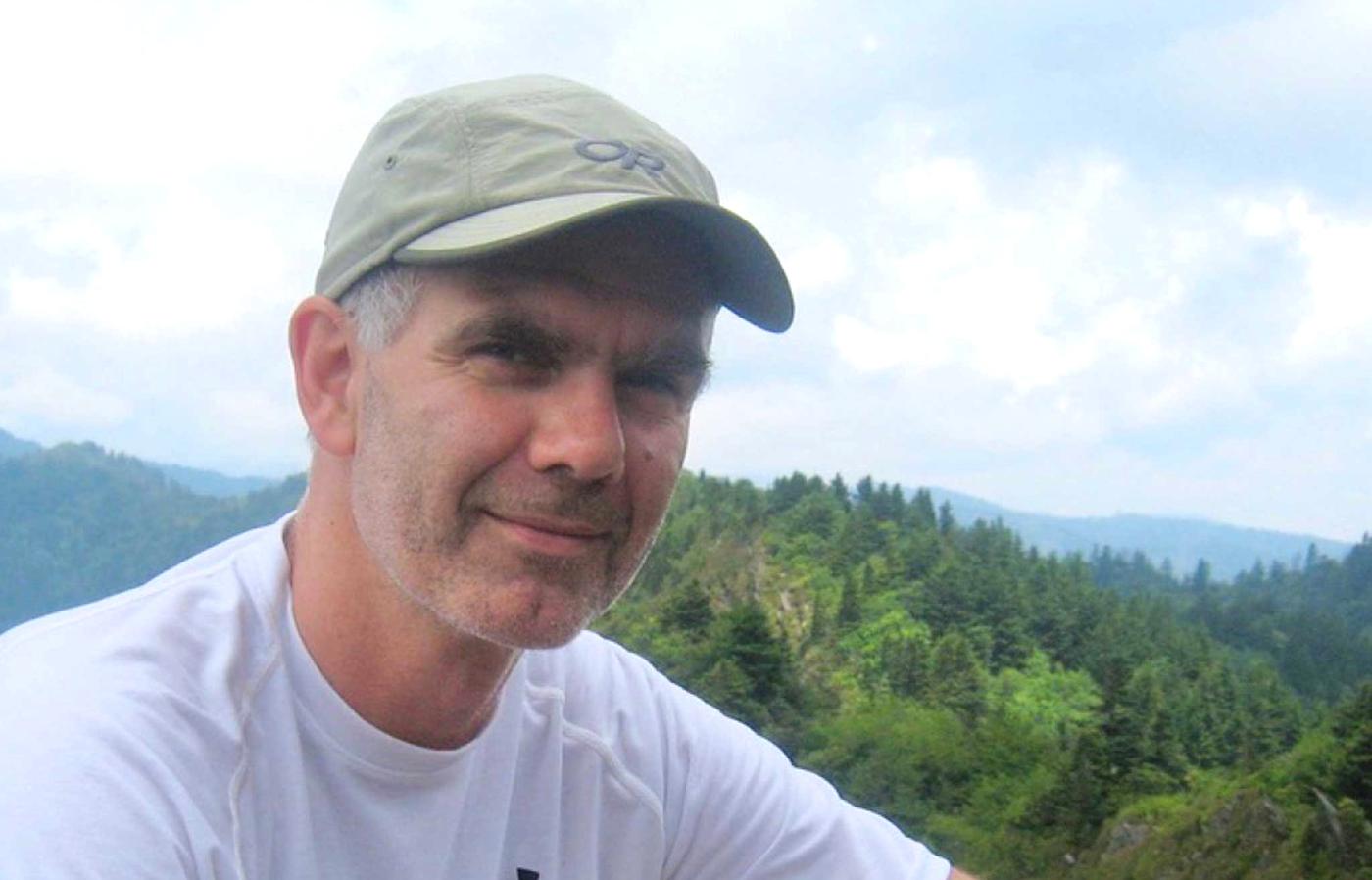 A person in a baseball cap sitting outside at a high vantage point with mountains in the background