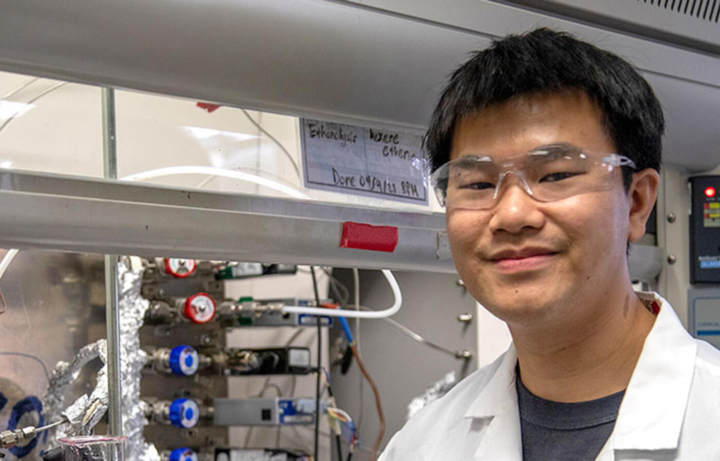 A person wearing a lab coat and eye protection stands in front of equipment in a lab facing the camera.