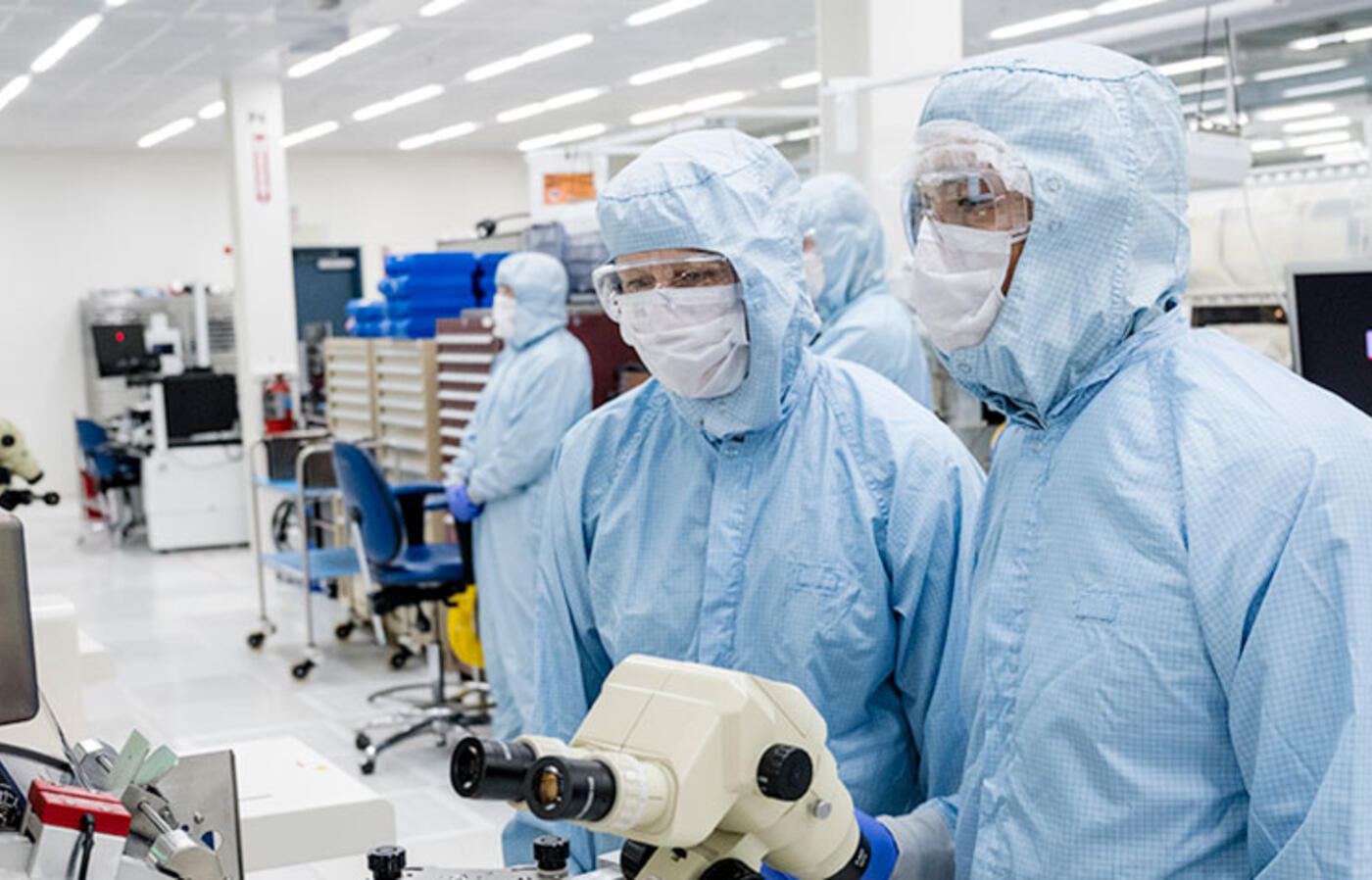 Two people wear cleanroom suits and stand in front of a microscope. 