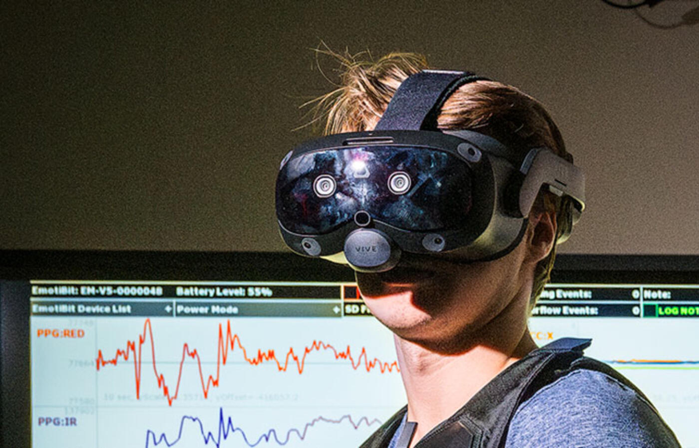 A person wears an augmented reality headset inside a lab and a digital screen displays data in the background.