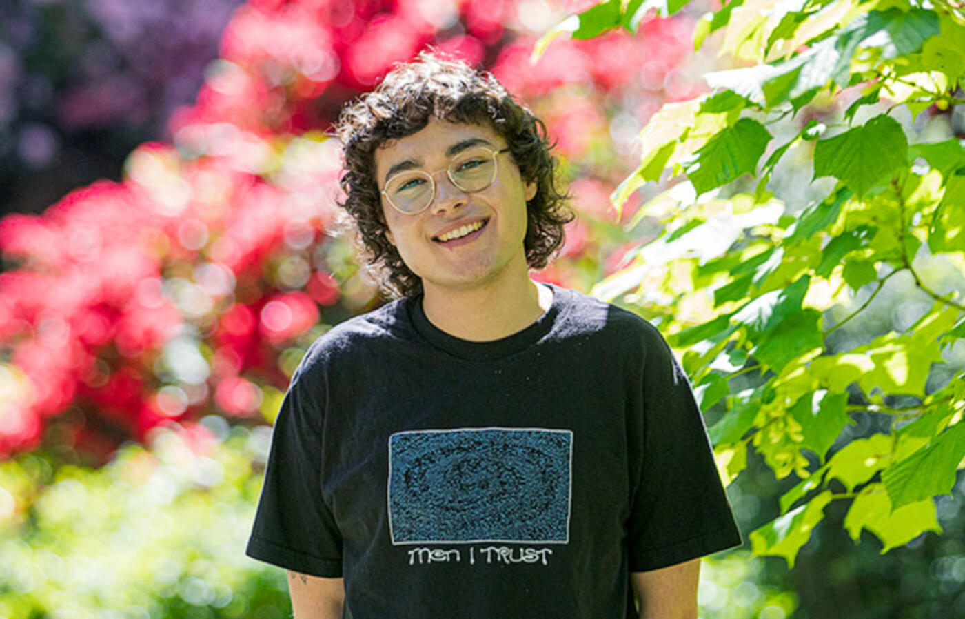 A person wearing a t-shirt smiles at the camera and stands in front of trees and spring blooms.