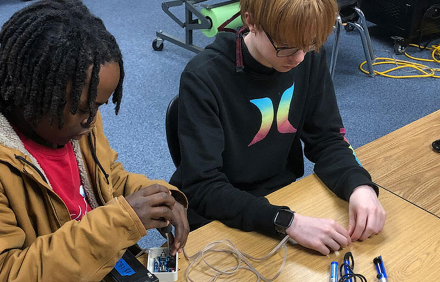 Two students sit at a table and adjust robotics equipment. 