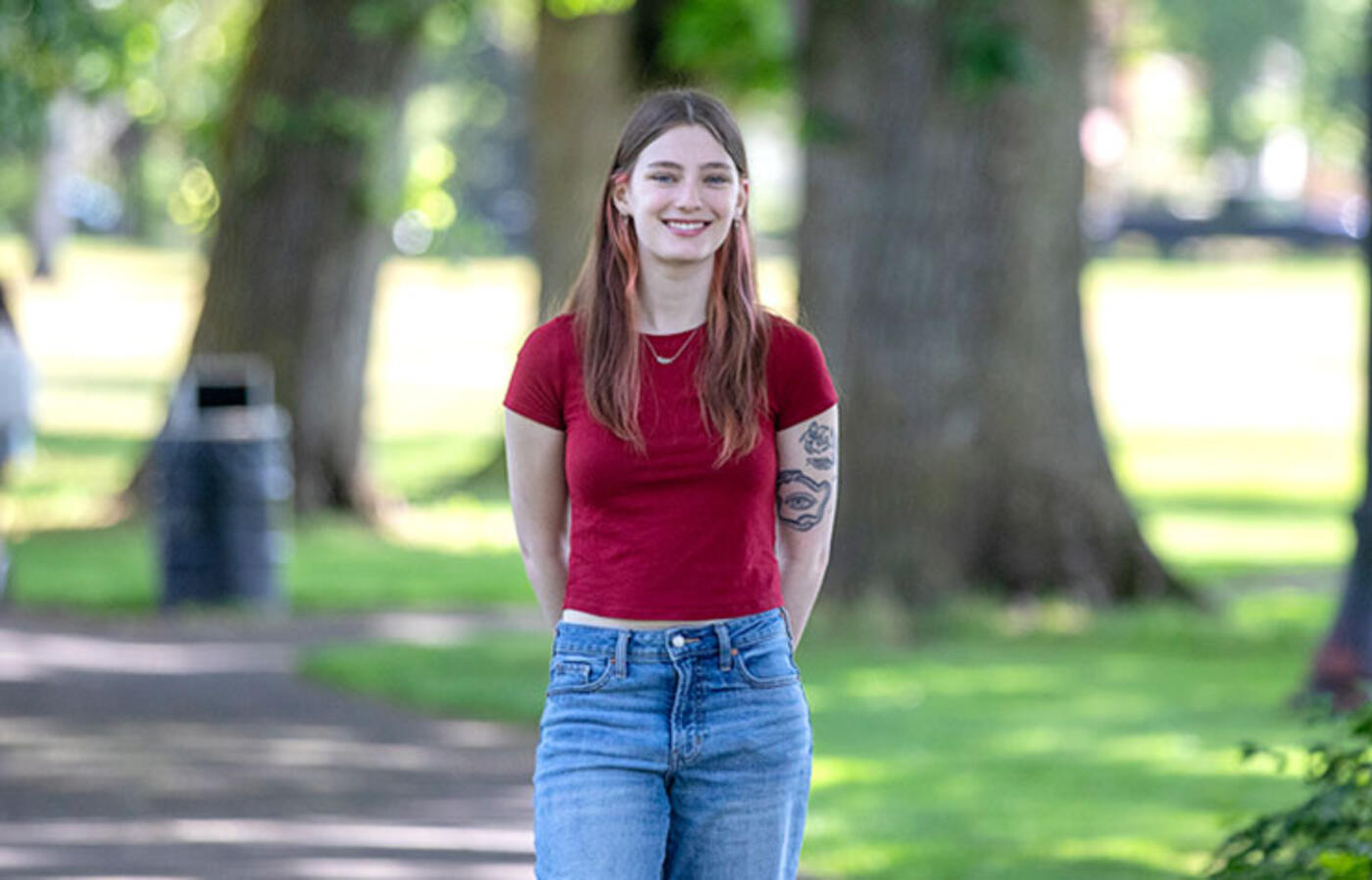 A person stands on a tree-lined path and smiles toward the camera. 