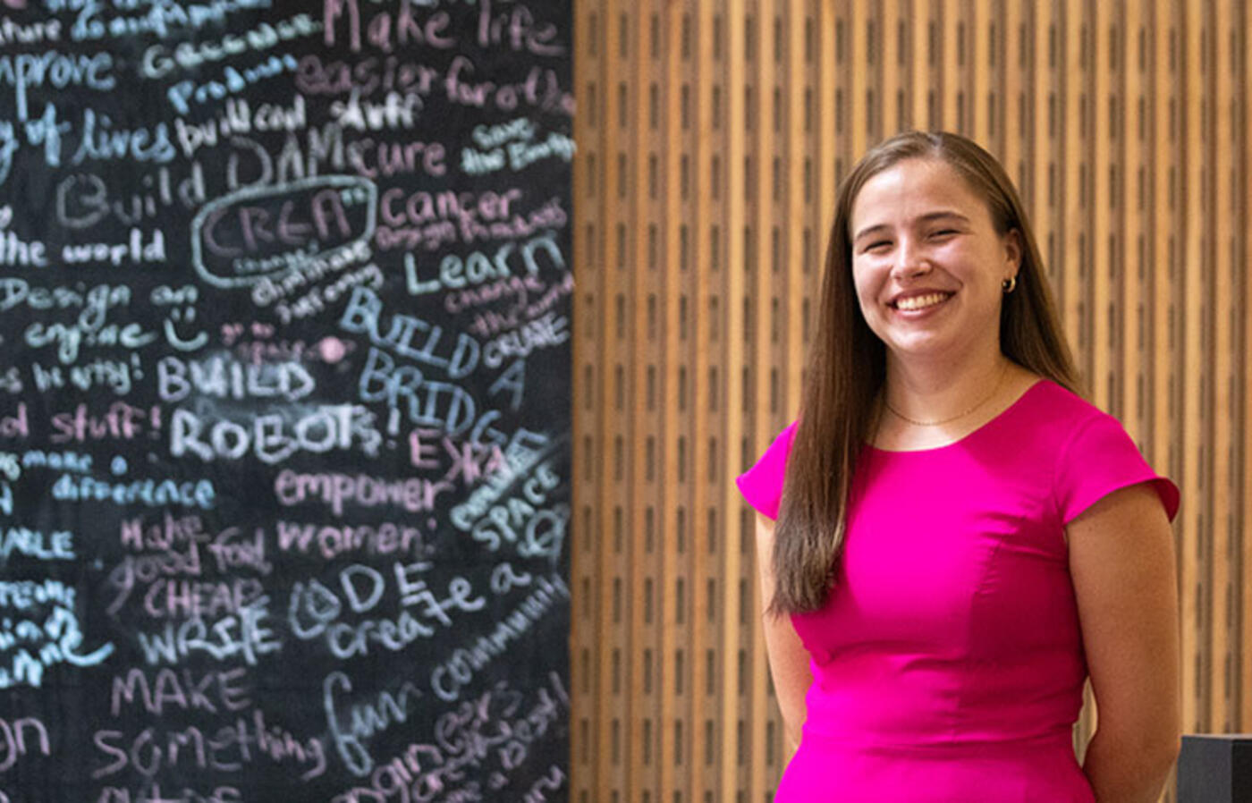 A person stands in front of a chalkboard with engineering terms written on it and smiles toward the camera.